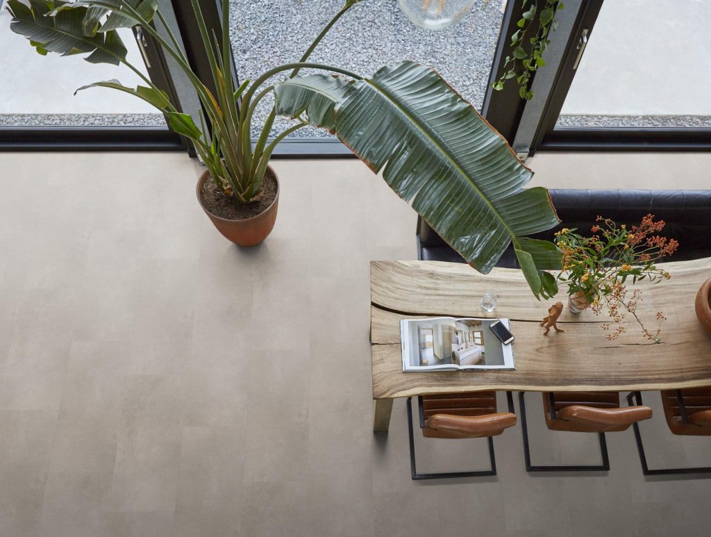 Bovenaanzicht van een eettafel van natuurlijk hout met leren stoelen op een lichte betonlook PVC vloer, naast een groot raam en een grote plant.