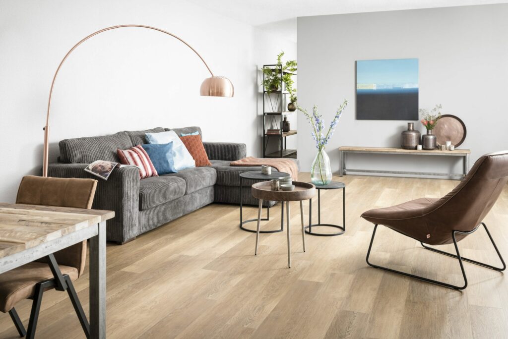 Moderne woonkamer met lichte houten vloer, grijze hoekbank, leren fauteuil, ronde salontafels en een booglamp in een rustige, neutrale inrichting.